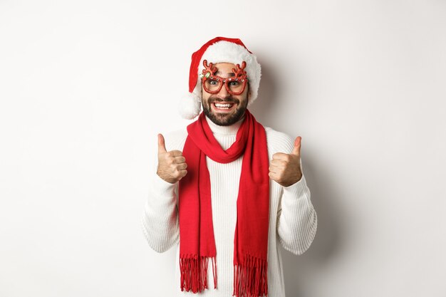 Natale, Capodanno e concetto di celebrazione. Uomo eccitato con cappello da Babbo Natale e occhiali da festa, che mostra i pollici in su in segno di approvazione, sorride soddisfatto, sfondo bianco