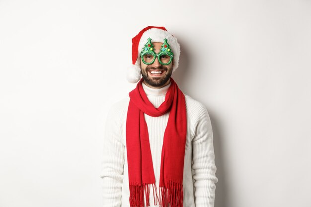 Natale, Capodanno e concetto di celebrazione. Uomo felice che ride, indossa cappello da Babbo Natale e occhiali da festa, in piedi su sfondo bianco