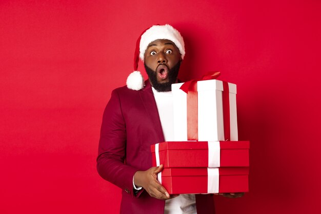 Natale, Capodanno e concetto di shopping. Allegro uomo nero segreto santa che tiene regali di Natale e sorride eccitato, porta regali, in piedi su sfondo rosso