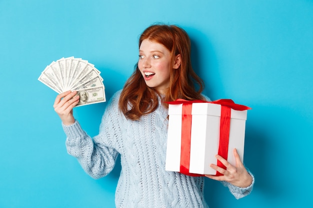 Natale e concetto di acquisto. Eccitata ragazza rossa che guarda i dollari, tiene in mano un grande regalo di Capodanno, compra regali, sta in piedi su sfondo blu