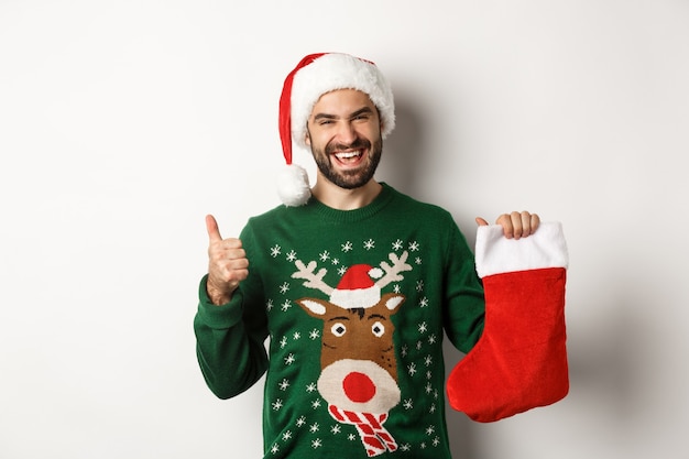 Natale e concetto di vacanze invernali. Uomo felice e contento con il cappello di Babbo Natale, che gradisce il regalo nel calzino di Natale, che mostra il pollice in su, in piedi su uno sfondo bianco