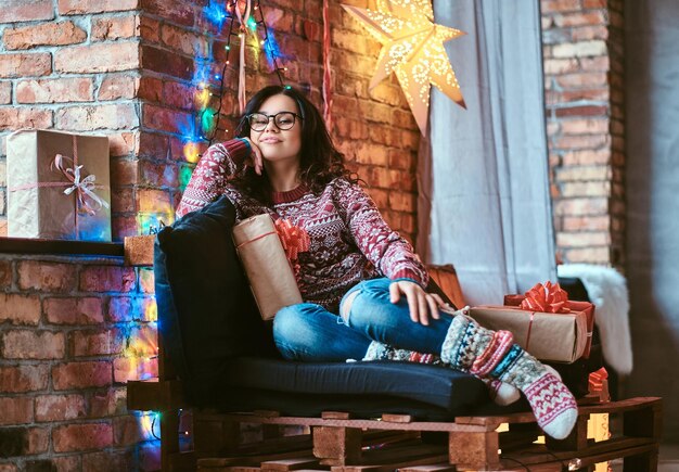 Natale, San Valentino, Capodanno. Felice bella ragazza godendo la mattina di Natale seduti su un divano con scatole regalo in una stanza decorata con interni soppalcati.