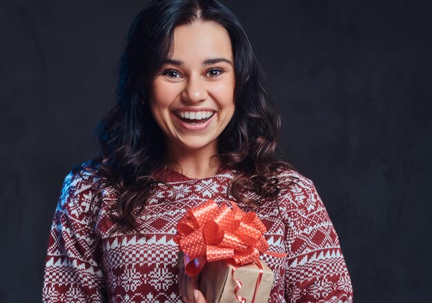 Natale, San Valentino, Capodanno. Ritratto di una ragazza bruna felice che indossa un maglione caldo con in mano una confezione regalo, isolata su uno sfondo scuro con texture.