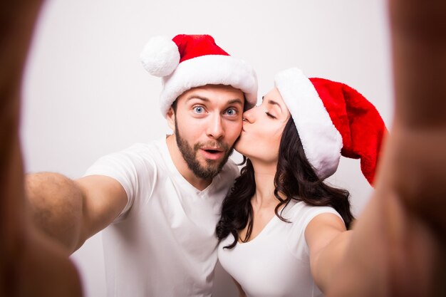 Natale, vacanze, tecnologia e concetto di persone - coppia felice in cappelli di Babbo Natale che scattano foto selfie dalle mani su sfondo bianco. Guardano la telecamera e si baciano