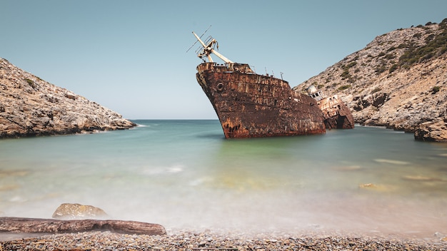 Nave arrugginita abbandonata nel mare vicino alle formazioni rocciose enormi sotto il chiaro cielo