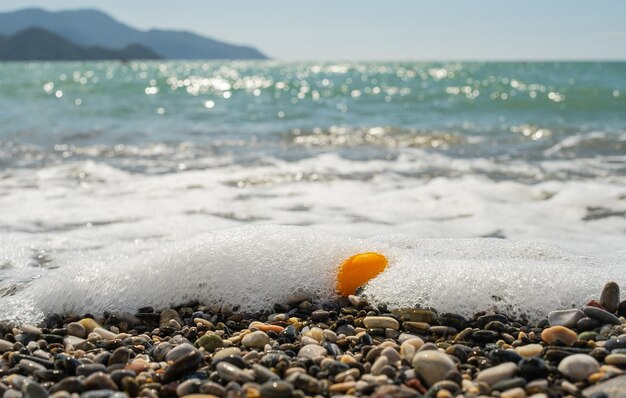 Navigare su una spiaggia di ciottoli un frutto di kumquat arancione brillante in schiuma di mare portato da un'onda alla spiaggia messa a fuoco selettiva sul mare in una giornata di sole luminosa tempo di vacanza o idea per uno sfondo