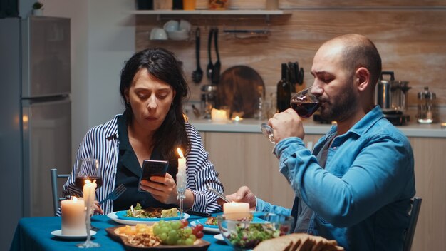 Navigare sui telefoni durante la cena coppia che tiene gli smartphone in cucina seduti al tavolo navigando, cercando, usando smartphone, internet, festeggiando il loro anniversario nella sala da pranzo.