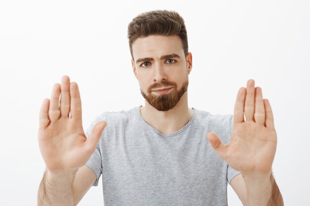 Ne ho abbastanza. Ritratto di uomo caucasico bello e maschile sotto pressione e dispiaciuto con barba e occhi azzurri che solleva i palmi nel gesto di arresto sorridendo per antipatia dando rifiuto sopra il muro grigio