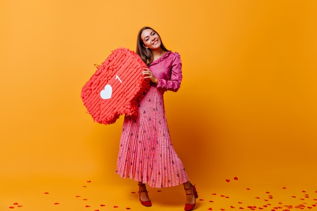 Nella stanza con la parete di cartone arancione c'è un'elegante donna dai capelli castani in gonna lunga, con un grande cartello da Instagram. Foto scattata nei colori arancio e rosa