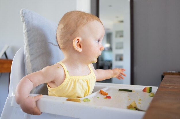 Neonata dai capelli rossi carina con macchie di purea verde sul viso seduto nel seggiolone con cibo disordinato sul vassoio e distogliere lo sguardo. Vista laterale. Processo di alimentazione o concetto di assistenza all'infanzia