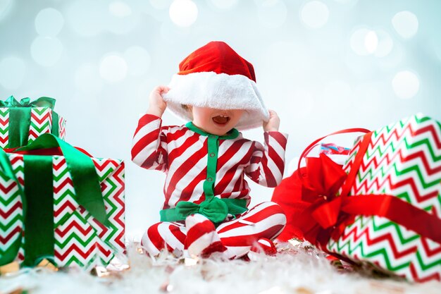 Neonata sveglia che porta il cappello della Santa che posa sopra le decorazioni di Natale con i regali. Seduto sul pavimento con la palla di Natale. Stagione delle vacanze.