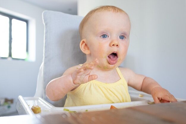 Neonata sveglia con le macchie di purea sul viso seduto nel seggiolone con cibo disordinato sul vassoio, aprendo la bocca e mostrando la lingua. Riflesso gargarismi o concetto di cura del bambino