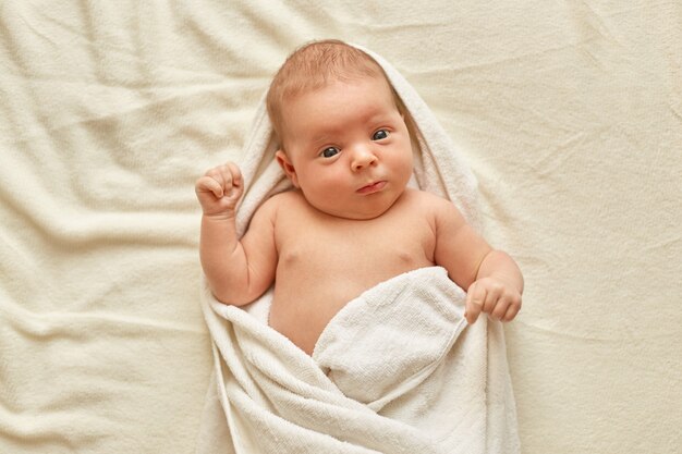 Neonato che indossa un asciugamano dopo il bagno, carino piccolo bambino sdraiato sul letto sulla coperta bianca, guardando e studiando le cose esteriori. Infanzia e cura del bambino.