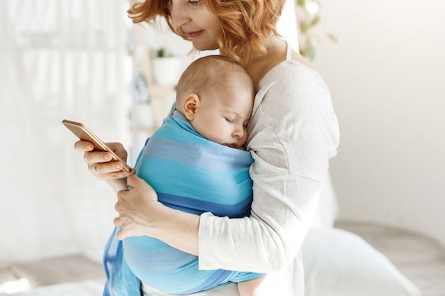 Neonato minuscolo che ha sogni piacevoli in imbracatura del bambino mentre la madre riposa e guardando attraverso i social network sullo smartphone. Famiglia, concetto di lifestyle.