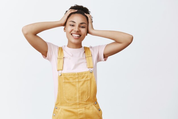 Nessuno stress, tempo per il relax. Ritratto di spensierata bella ragazza afroamericana felice in tuta elegante gialla, toccando i capelli e sorridendo ampiamente, essendo di ottimo umore dopo un ottimo pisolino pranzo