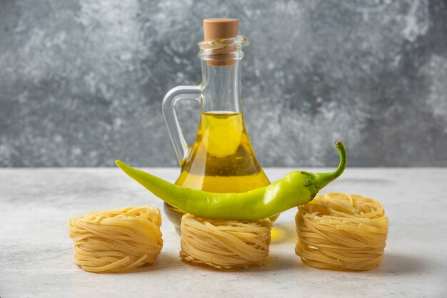 Nidi di pasta cruda, bottiglia di olio d'oliva e pepe verde sul tavolo bianco.