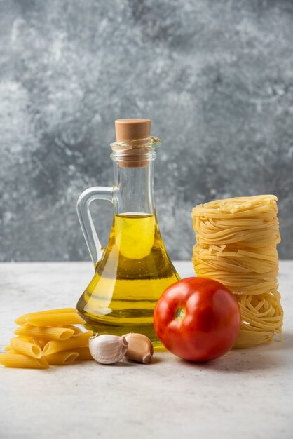 Nidi di pasta cruda, bottiglia di olio d'oliva e verdure sulla tavola bianca.