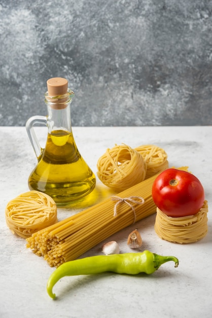 Nidi di pasta cruda, spaghetti, bottiglia di olio d'oliva e verdure su superficie bianca.