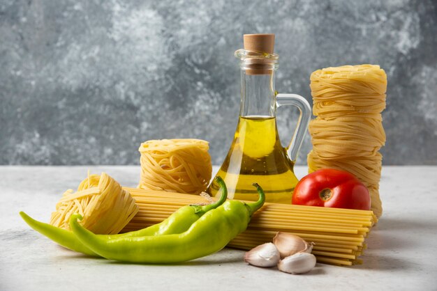 Nidi di pasta cruda, spaghetti, bottiglia di olio d'oliva e verdure su superficie bianca.