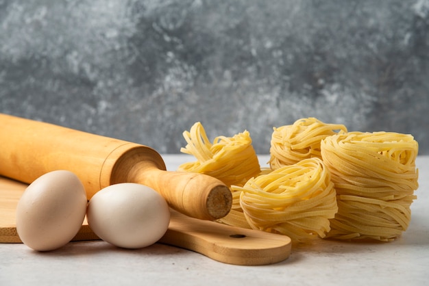 Nidi di pasta cruda, uova, tavola di legno e mattarello sul tavolo bianco.