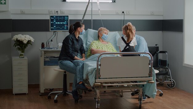 Nipote in visita a un paziente anziano con maschera facciale in corsia d'ospedale con madre e vecchio. Donna malata che si gode la visita di figlia e figlio, dando conforto per curare la malattia durante la pandemia.