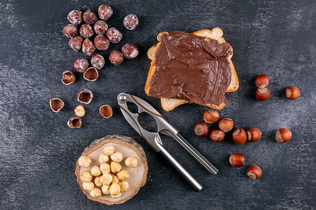 Nocciole sgusciate con pane spalmato di cacao, schiaccianoci, pezzo di legno vista dall'alto su un tavolo di pietra scura