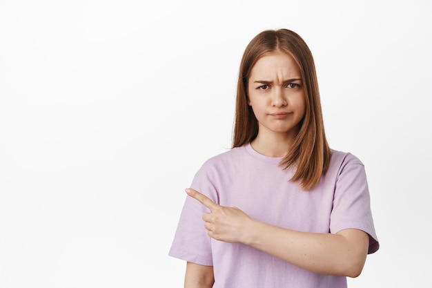 Non bene. Ritratto di ragazza bionda solca le sopracciglia da antipatia, puntando il dito a sinistra e lamentandosi, delusa da qualcosa di cattivo, condanna, in piedi su sfondo bianco.