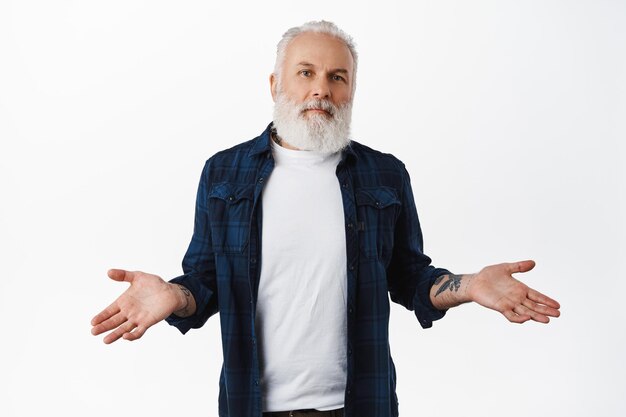 Non lo so. Uomo anziano incapace con tatuaggi, alzando le spalle e guardando confuso, perplesso o inconsapevole, non ho idea, non capisco, in piedi su sfondo bianco