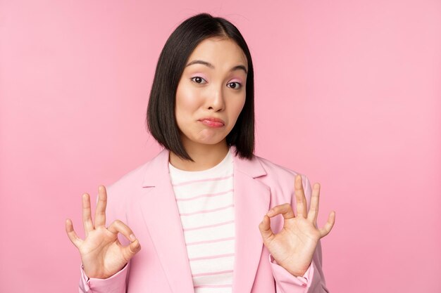 Non male Impressionato donna d'affari asiatica dell'ufficio che mostra segno ok e cenno di approvazione in piedi su sfondo rosa Spazio di copia