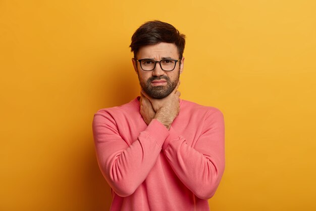 Non riesco a respirare. Il ragazzo unshven frustrato e angosciato tiene le mani sul collo, soffoca e sente mal di gola, ha attacchi asmatici, sente disagio e sensazioni dolorose, posa al chiuso, ha bisogno di antidolorifici