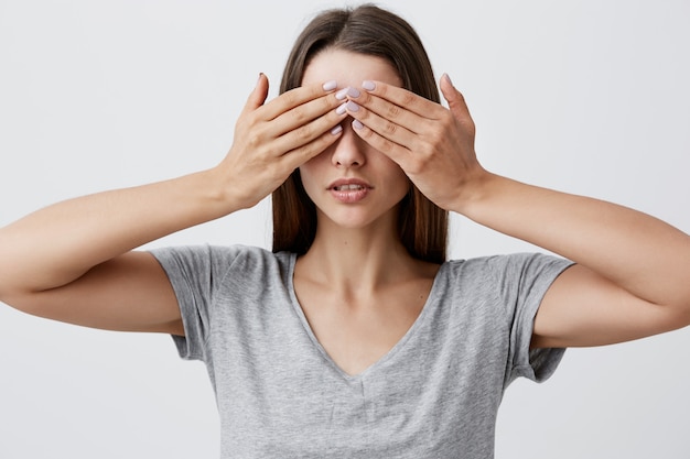 Non vedo niente Il ritratto di giovane bella donna caucasica sexy dell'allievo con capelli lunghi scuri in vestiti grigi della maglietta osserva con le mani con espressione seria e calma del fronte. Linguaggio del corpo.