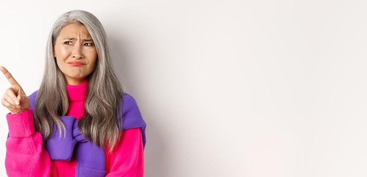 Foto gratuito nonna asiatica dispiaciuta in maglione rosa che fa smorfie e accigliata scettica puntando il dito in alto
