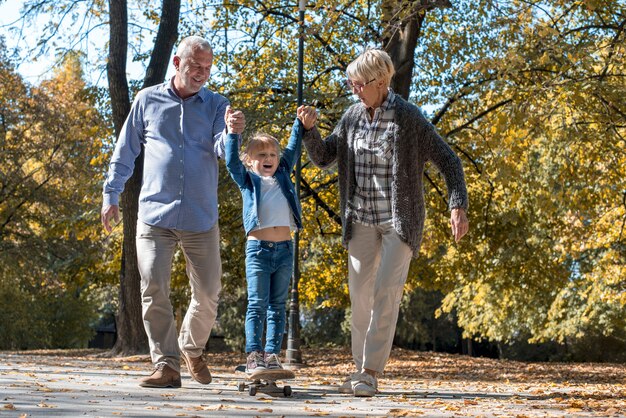 Nonni sorridenti che tengono le mani della nipote mentre sta cavalcando lo skateboard in un parco autunnale