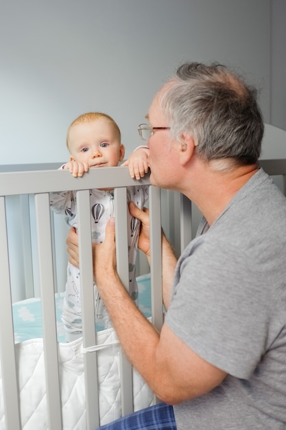 Nonno che prepara il bambino sveglio a stare in piedi. Bambino in piedi nel presepe e guardando la fotocamera. Colpo verticale. La cura dei bambini o il concetto di infanzia