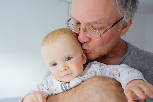 Nonno che tiene in braccio e che bacia il bambino sveglio dagli occhi blu. Colpo del primo piano. La cura dei bambini o il concetto di infanzia