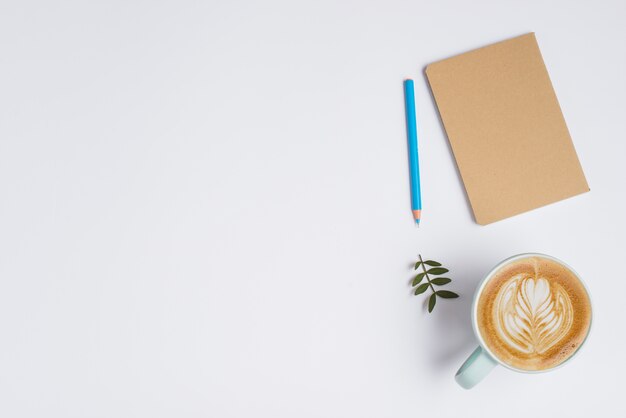 Notebook chiuso; matita colorata; foglie e tazza di caffè con latte art su sfondo bianco