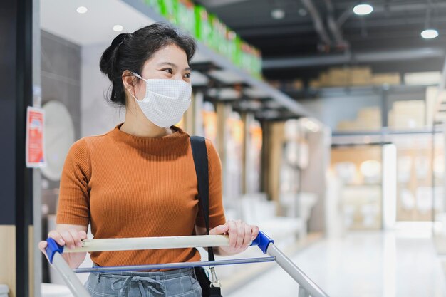 Nuovo stile di vita normale asiatico femminile abito marrone indossare maschera protettiva shopping nei grandi magazzini dopo il periodo di quarantena di blocco Nuovo stile di vita normale uso mano smartphone bokeh sfondo