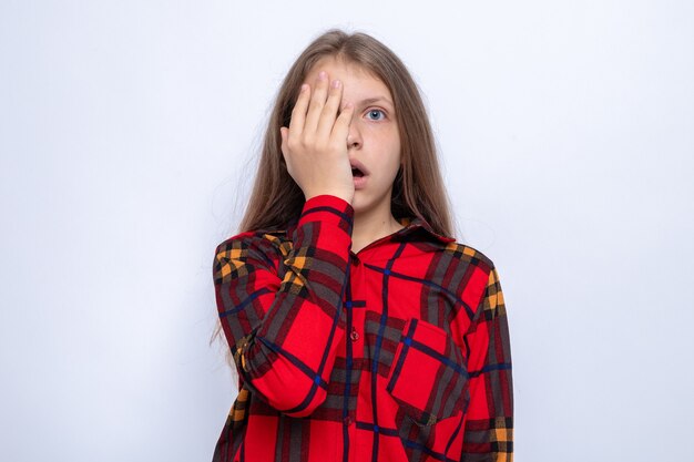 Occhi coperti spaventati con la mano bella bambina che indossa una camicia rossa isolata sul muro bianco