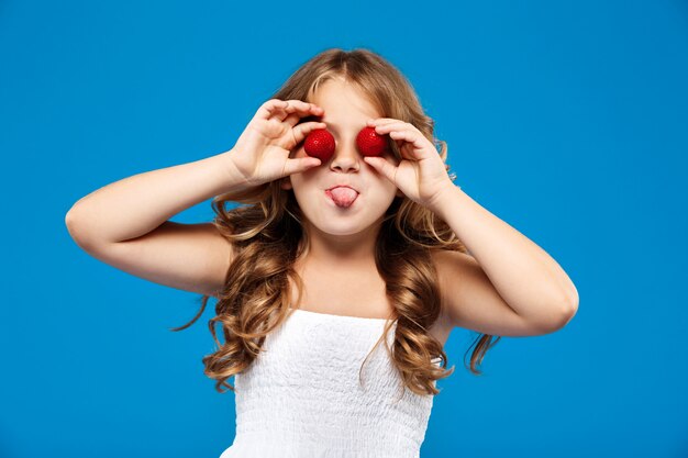 Occhi nascondentesi della giovane ragazza graziosa con la fragola sopra la parete blu