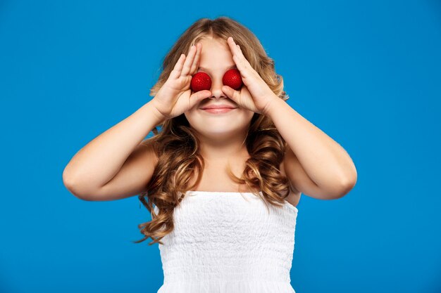 Occhi nascondentesi della giovane ragazza graziosa con la fragola sopra la parete blu