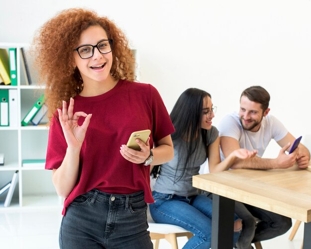 Occhiali da portare della donna graziosa che gesturing segno giusto con i suoi amici che discutono dietro