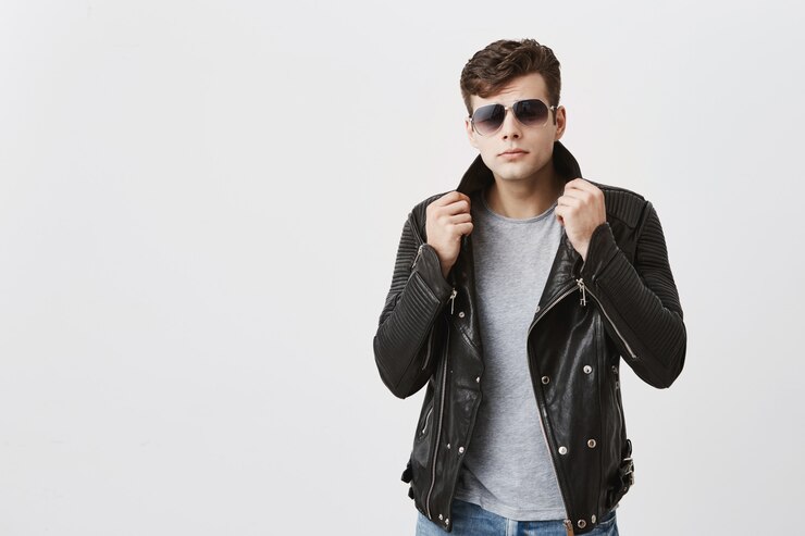 Foto gratuito occhiali da sole da portare del maschio sicuro muscolare che propongono all'interno. bel ragazzo attraente con taglio di capelli alla moda in giacca di pelle nera e jeans, tirando con le mani giacca in su, guardando con appello