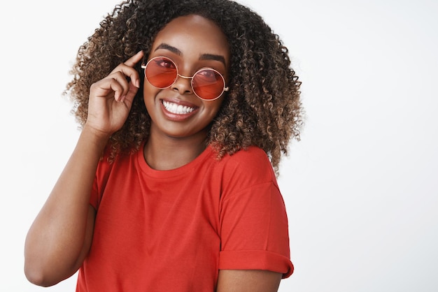 Occhiali da sole luminosi per qualsiasi condizione atmosferica Ritratto di affascinante spensierata e carismatica bella donna afroamericana in maglietta rossa e occhiali da sole alla moda che sorride ampiamente alla fotocamera sul muro bianco