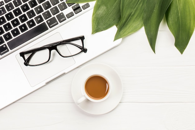 Occhiali e foglie neri su un computer portatile aperto con la tazza di caffè sullo scrittorio di legno