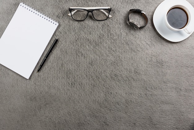 Occhiali; orologio da polso; tazza di caffè e blocco note a spirale con penna sul tavolo grigio