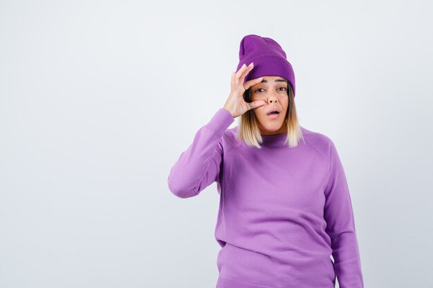 Occhio di apertura della giovane signora con il dito in maglione viola, berretto e guardando perplesso, vista frontale.