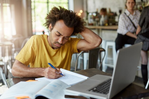 Occupato studente afroamericano seduto in caffetteria affrettandosi a scrivere note nel suo quaderno utilizzando il computer portatile per la ricerca di informazioni grattandosi la testa con la mano. Istruzione, concetto di gioventù