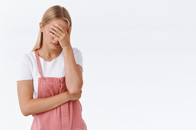 Oddio perché io. Giovane ragazza bionda infastidita imbarazzata e stanca, fai facepalm, tieni la mano sul viso, cercando di calmarti come irritata e incazzata, in piedi a disagio su sfondo bianco