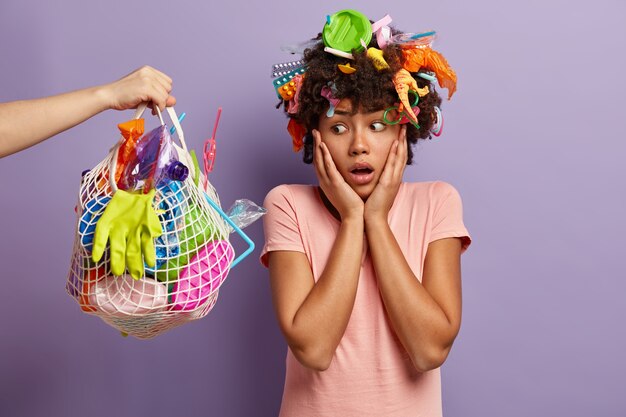 Oh no, non inquinare la natura con i rifiuti di plastica! La donna etnica infelice guarda con espressione scioccata al sacco pieno di immondizia di plastica, pulisce il pianeta, pone al coperto. Giornata della Terra e concetto di volontariato