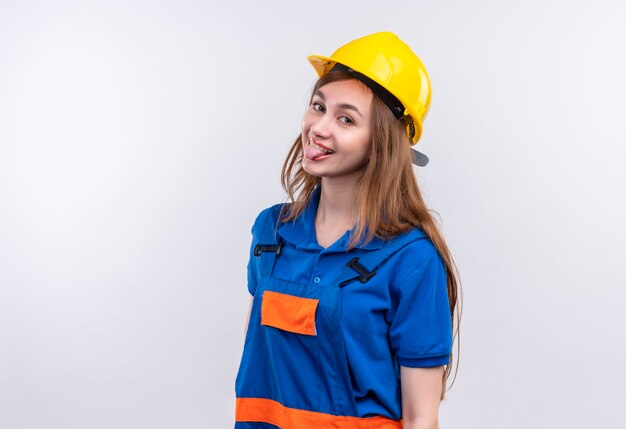 Operaio costruttore allegro giovane donna in uniforme da costruzione e casco di sicurezza che attacca fuori la lingua